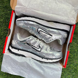 A pair of Nike running shoes in metallic silver and white, displayed inside an open shoebox lined with white tissue paper, set on green grass.