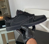 A black low-top sneaker with white contrast stitching and a black sole, held up by a hand wearing a black glove. The shoe has a black Nike Swoosh on the side and a black Nike tongue tag, with a small red 'X' detail on the tongue.